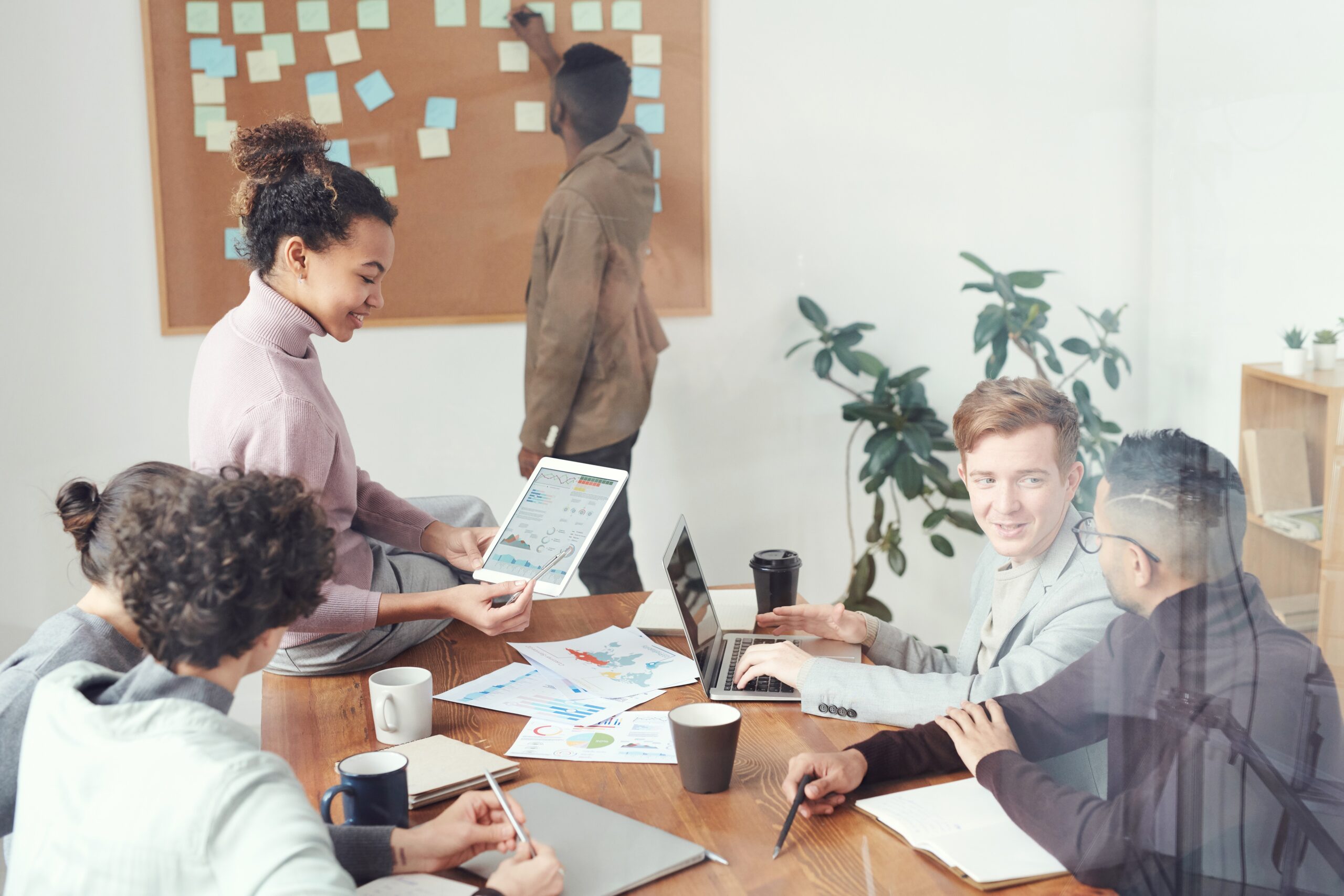 A number of people sitting around a table working together. Another peraon standing up adding post it notes to a cork board.